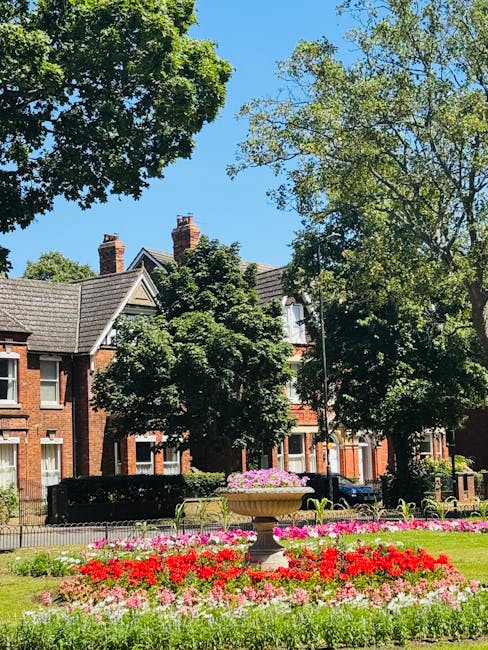 The image depicts a vibrant flower bed located in a park setting, with a large stone planter filled with pink and purple flowers positioned centrally on a well-maintained grassy area. Surrounding the