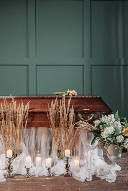 A funeral flower display featuring a large arrangement of white roses, lilies, and carnations in a white ceramic vase, complemented by sprigs of greenery. The flowers are fresh with vibrant petals and