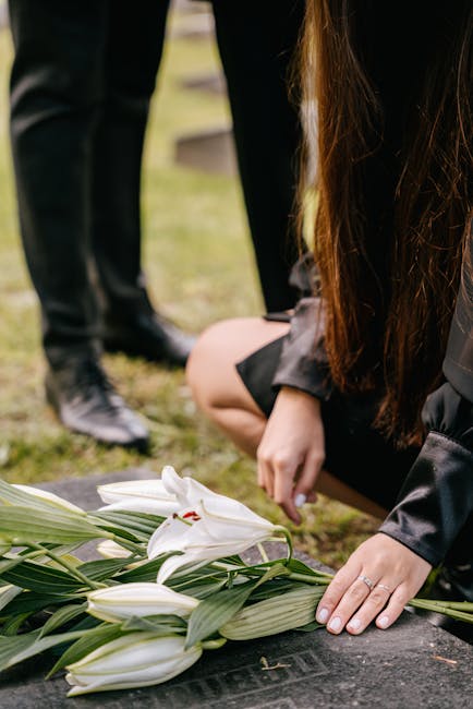 A person laying white lilies with large green leaves on a dark stone grave, gently adjusting the floral arrangement during a funeral service, with other people in dark clothing visible in the backgrou