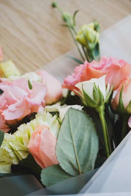 A close-up of a floral arrangement of fresh Lisianthus flowers with delicate, ruffled petals in shades of soft pink and creamy white, accompanied by green foliage with broad, smooth leaves. The bouque
