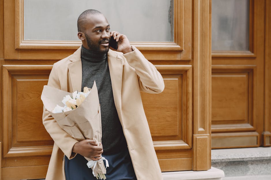 A man with a dark complexion and a neatly trimmed beard is standing outdoors in front of a wooden-paneled building, holding a bouquet of fresh white flowers wrapped in brown craft paper with some gree