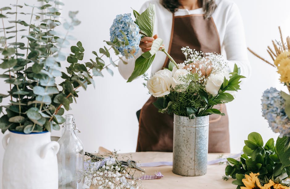A florist arranging a floral gift featuring white roses, blue hydrangeas, and sprigs of baby's breath in a tall, cylindrical, silver vase. The florist is wearing a white top and brown apron, holding t