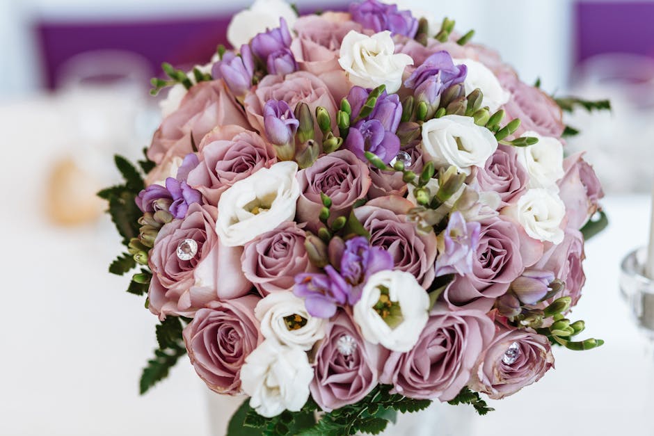 A close-up of a round floral bouquet featuring predominantly soft pink roses, accented with white and purple lisianthus, and small purple freesias, all arranged with lush green foliage. The bouquet is